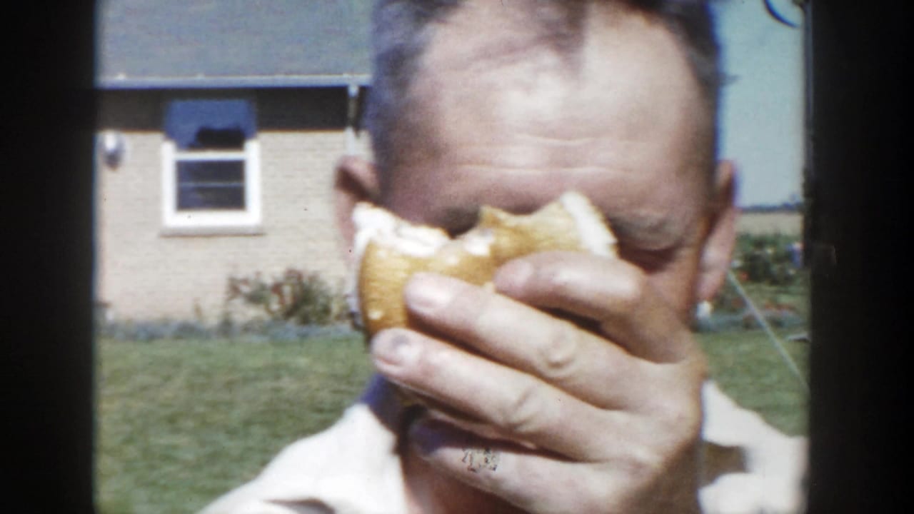 In 1958 Wisconsin home-movie footage, a man in a white collared shirt raises a sandwich near his face with brick houses and lawn behind him.