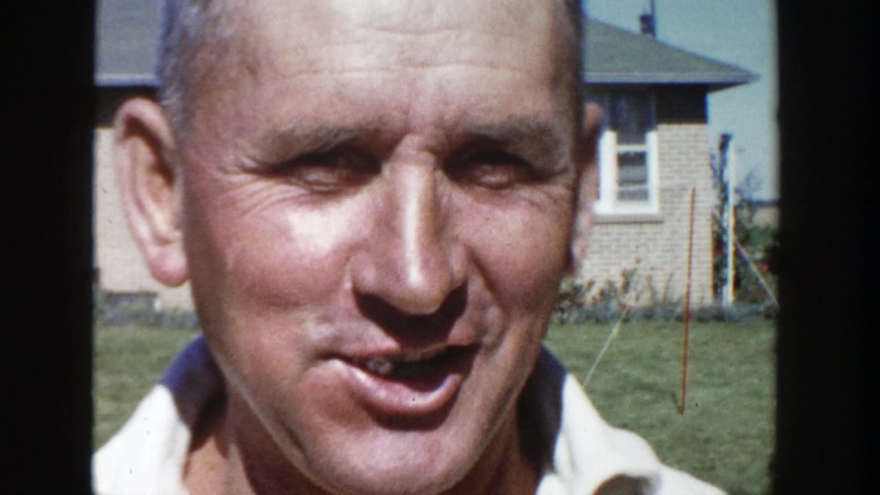 A close 1958 Wisconsin frame shows the man speaking toward the camera, with a brick house, lawn, and bright daylight in the background.