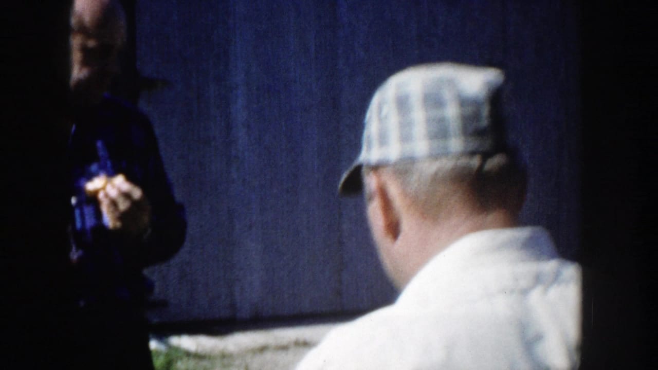 A 1958 Wisconsin home-movie frame shows the man from behind in a light shirt and plaid cap near a dark wall, with another person partly visible.