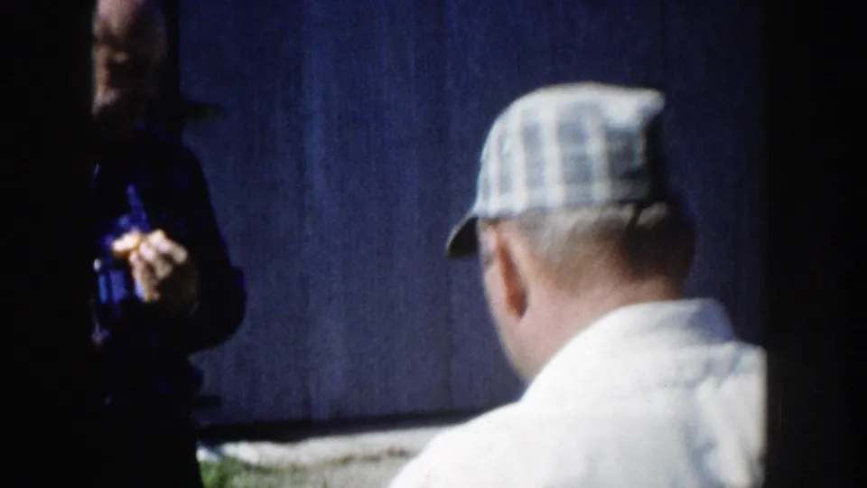 A 1958 Wisconsin home-movie frame shows the man from behind in a light shirt and plaid cap near a dark wall, with another person partly visible.