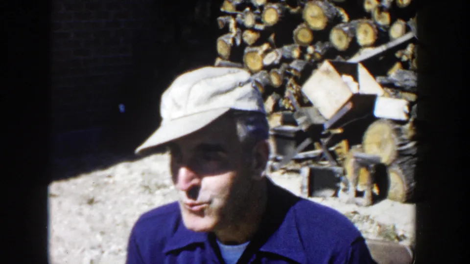 In 1958 Wisconsin home-movie footage, a man in a light cap and blue work shirt stands near a large stack of chopped firewood.