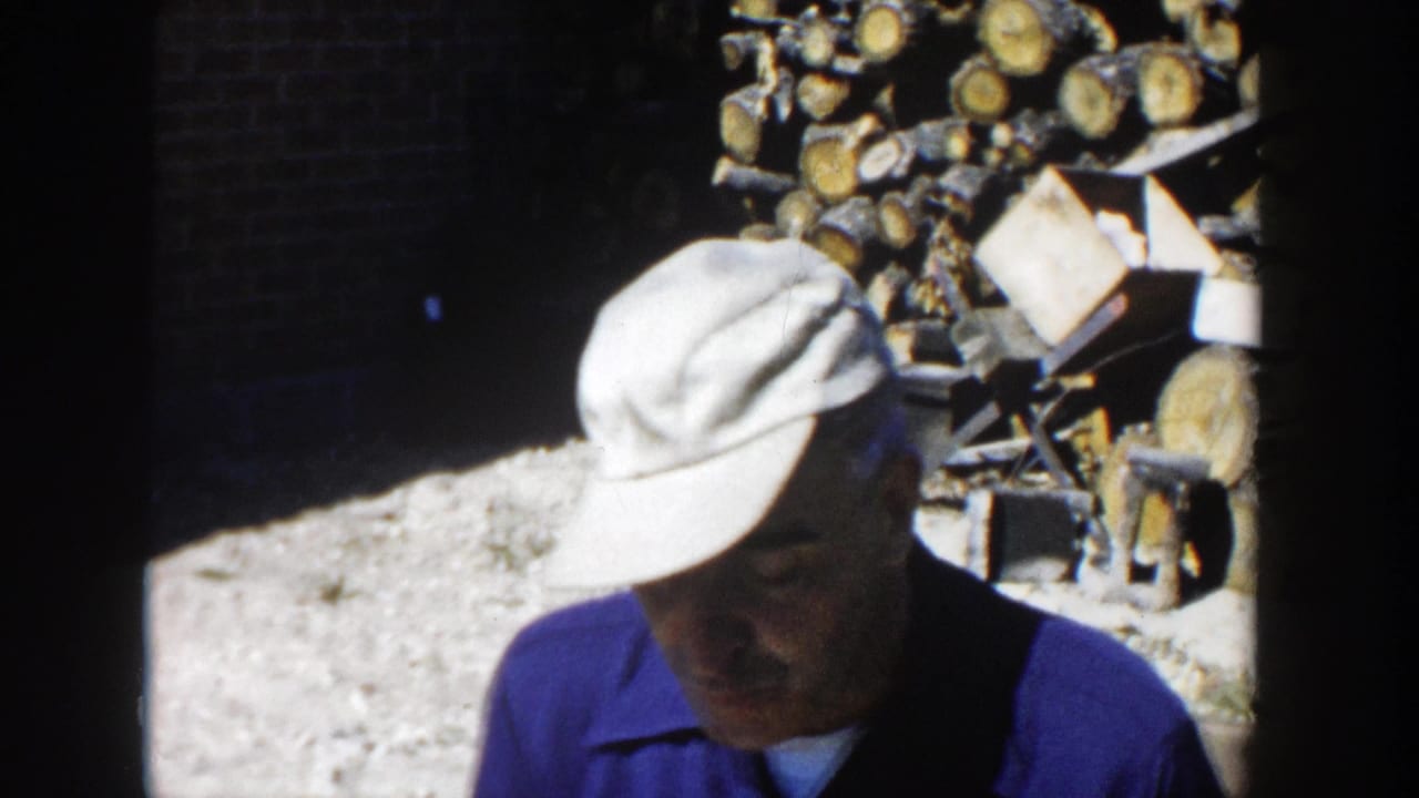 A man in a light cap and blue shirt looks down while standing close to a woodpile in the 1958 Wisconsin home-movie scene.
