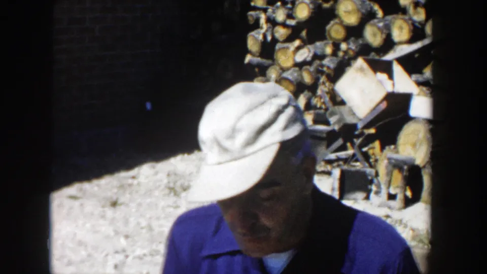A man in a light cap and blue shirt looks down while standing close to a woodpile in the 1958 Wisconsin home-movie scene.