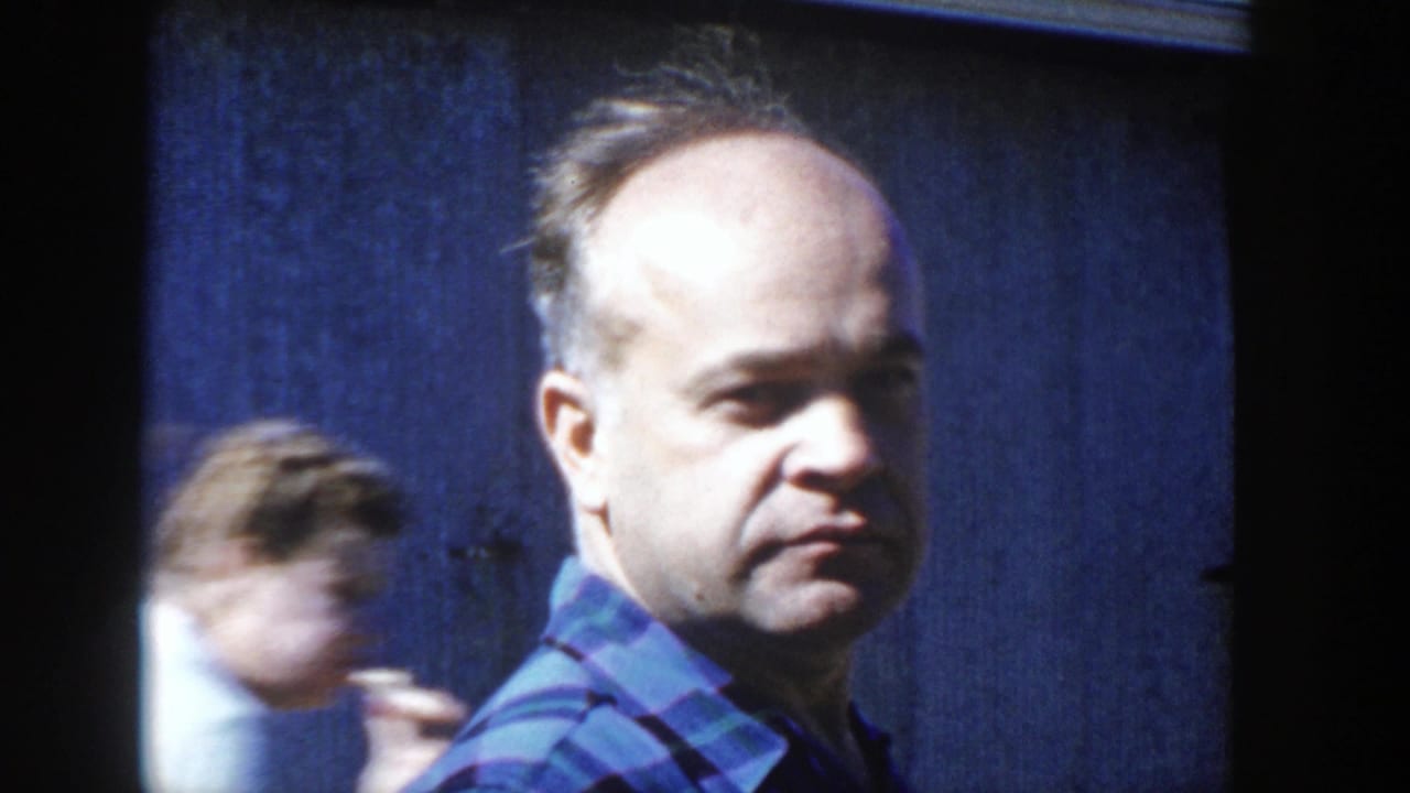 In Wisconsin 1958 home-movie footage, an adult man in a blue and green plaid shirt looks toward the camera beside a dark blue wall.