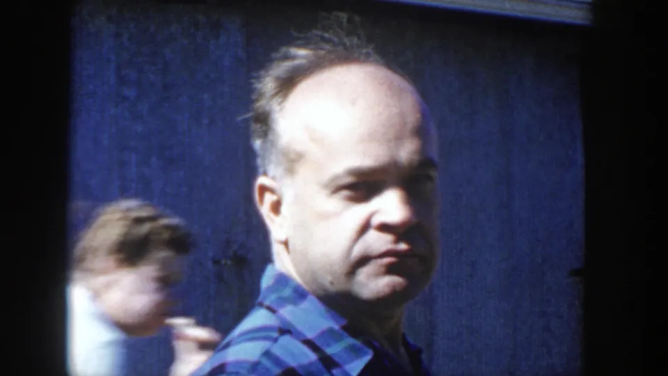 In Wisconsin 1958 home-movie footage, an adult man in a blue and green plaid shirt looks toward the camera beside a dark blue wall.