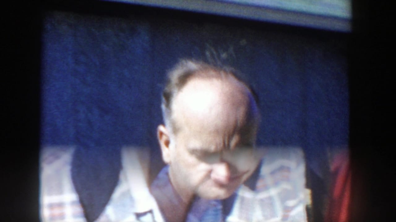 A close 1958 Wisconsin home-movie frame shows the plaid-shirted man looking down, with soft focus and dark wall behind him.