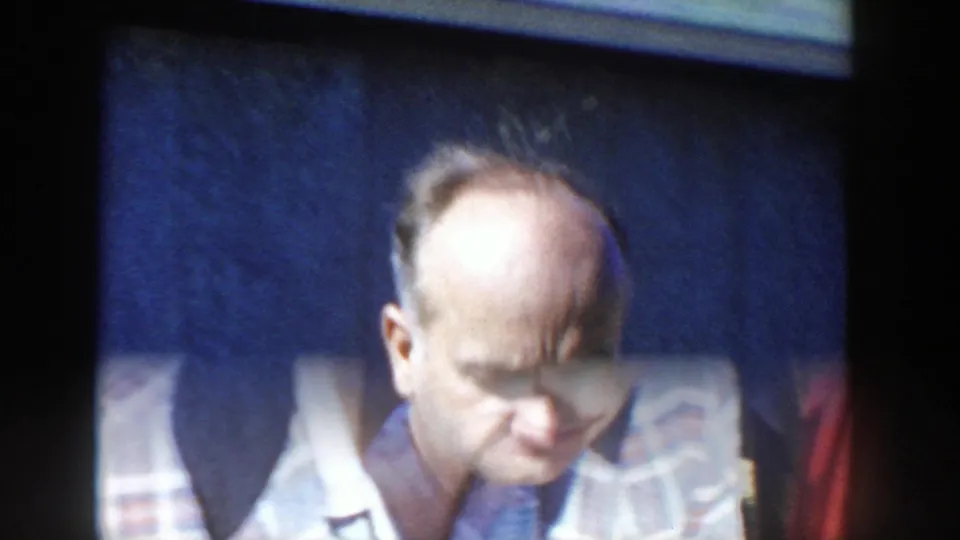 A close 1958 Wisconsin home-movie frame shows the plaid-shirted man looking down, with soft focus and dark wall behind him.