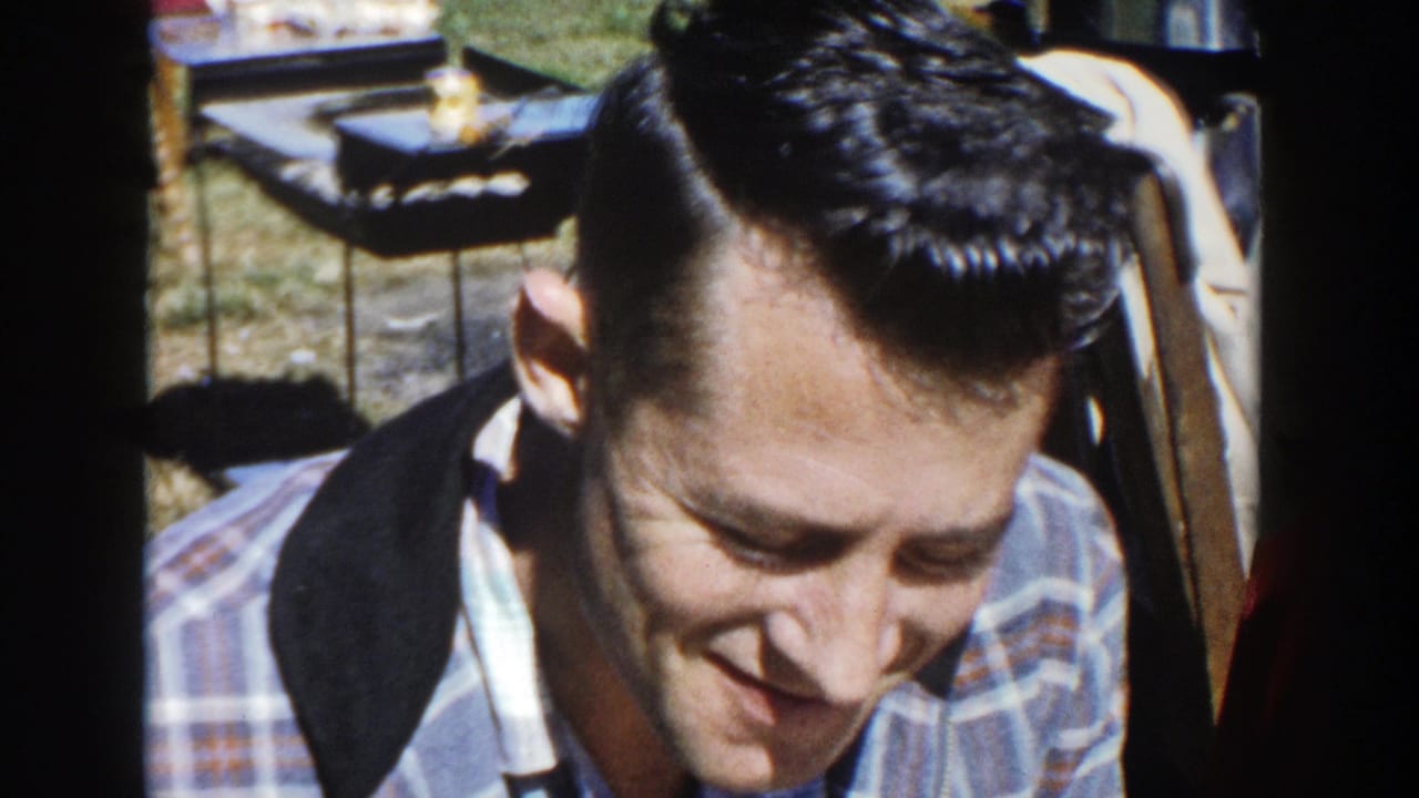A younger adult man with a pompadour and plaid shirt smiles while seated at a sunny picnic in Wisconsin 1958 footage.
