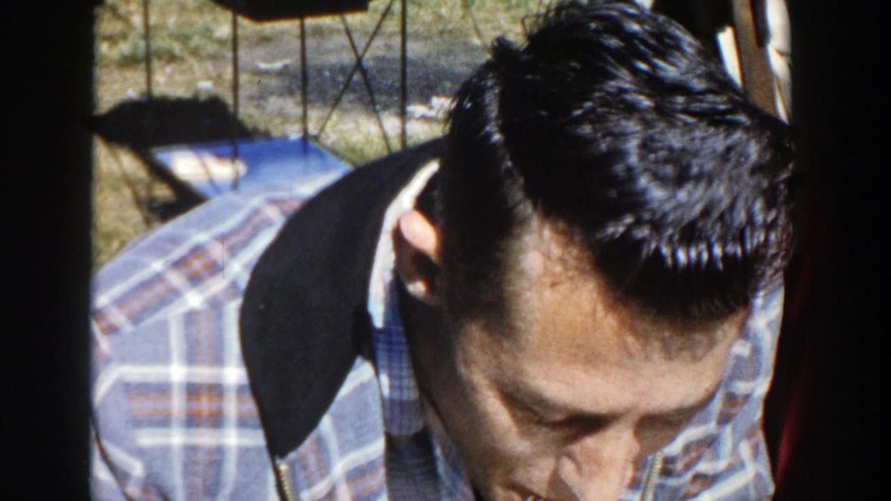 In supported 1958 Wisconsin home-movie footage, a young man in plaid sits close to camera at a picnic table in sunlight.
