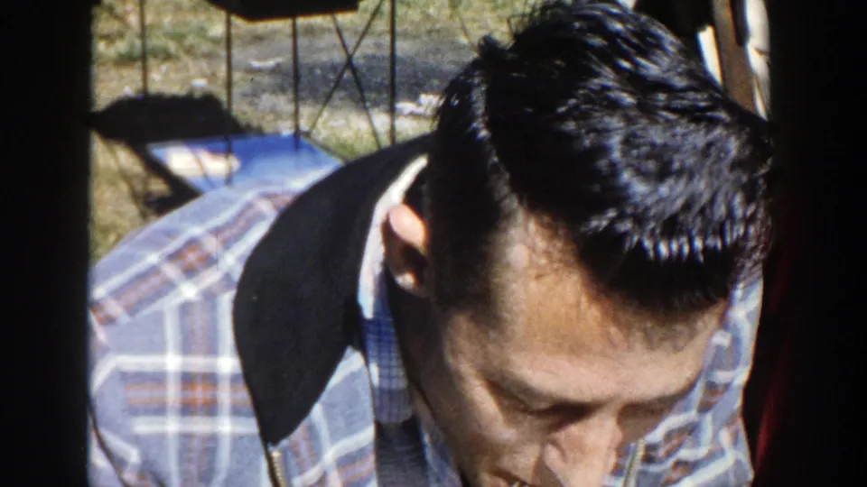 In supported 1958 Wisconsin home-movie footage, a young man in plaid sits close to camera at a picnic table in sunlight.