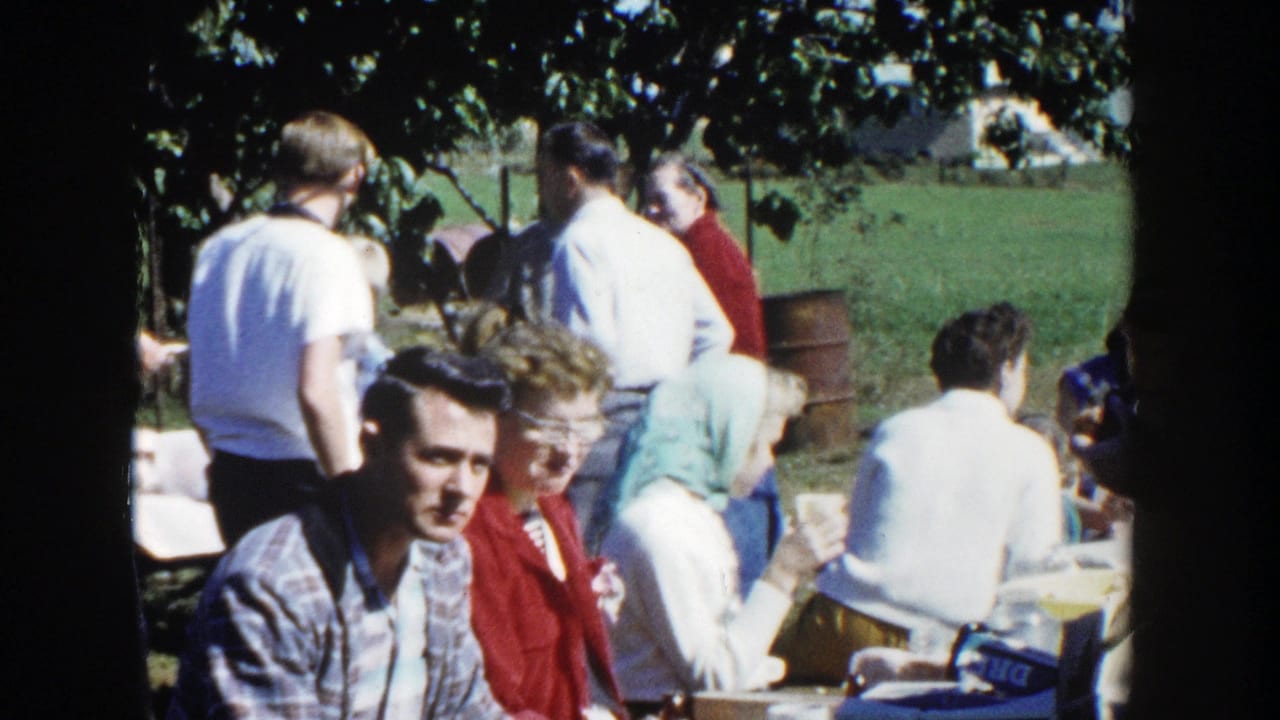 A wider home-movie view shows adults seated and standing under trees at a Wisconsin picnic, with cups and tables visible.