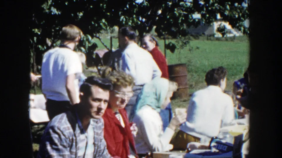 A wider home-movie view shows adults seated and standing under trees at a Wisconsin picnic, with cups and tables visible.