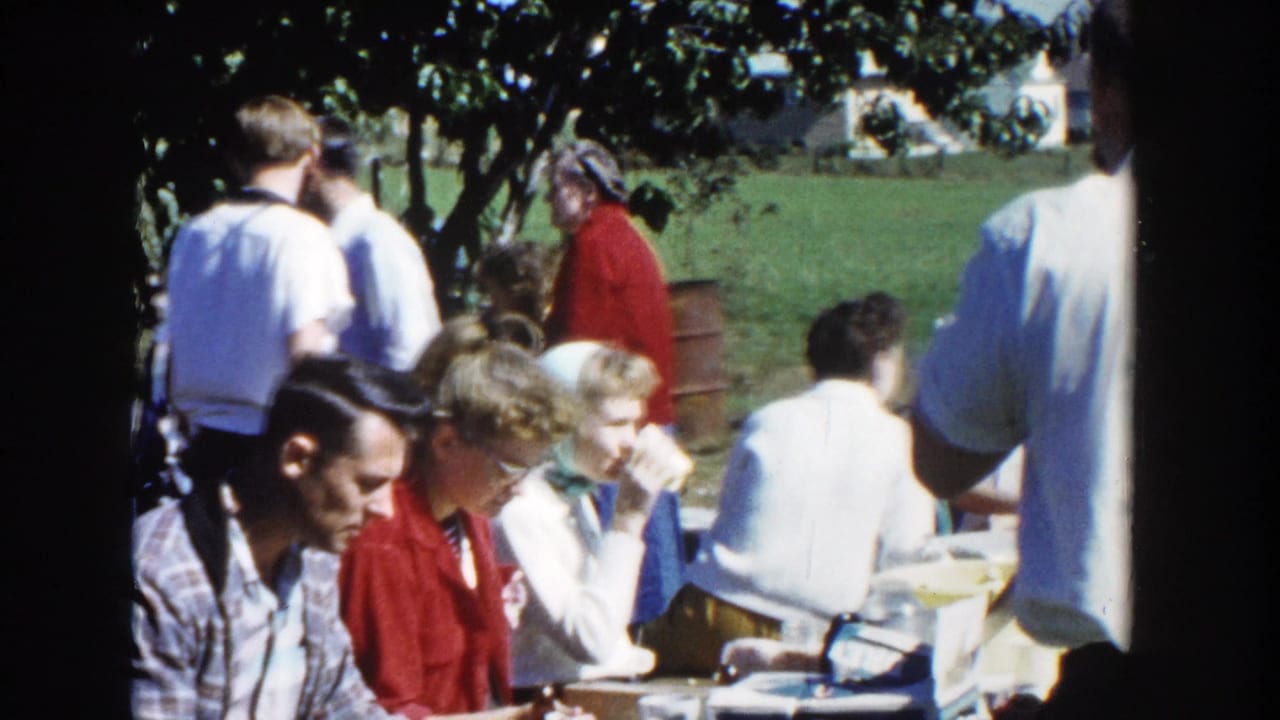 The clip ends on a wider 1958 Wisconsin picnic view with several adults eating and talking beneath trees in bright daylight.