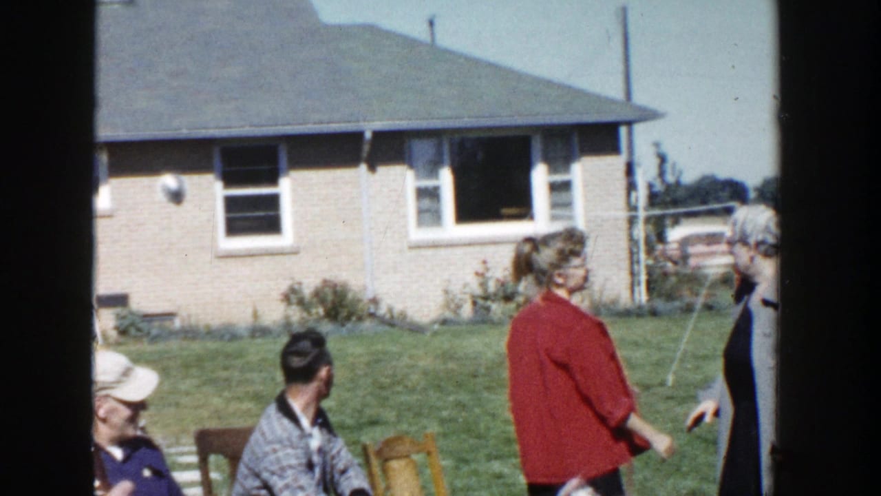 1958 Wisconsin home-movie frame of adults standing and sitting in a sunny backyard near a brick ranch house and stacked firewood.