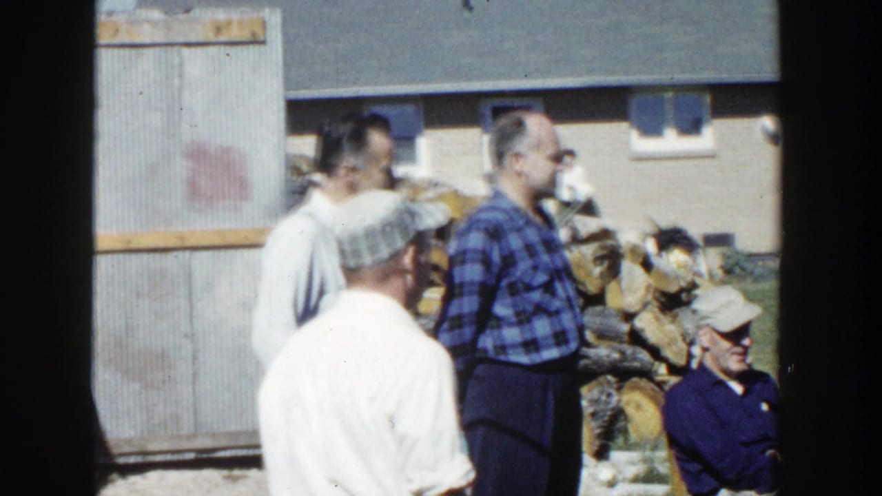 1958 Wisconsin home-movie frame showing adults beside a corrugated shed or fence, lawn chairs, and a backyard woodpile.