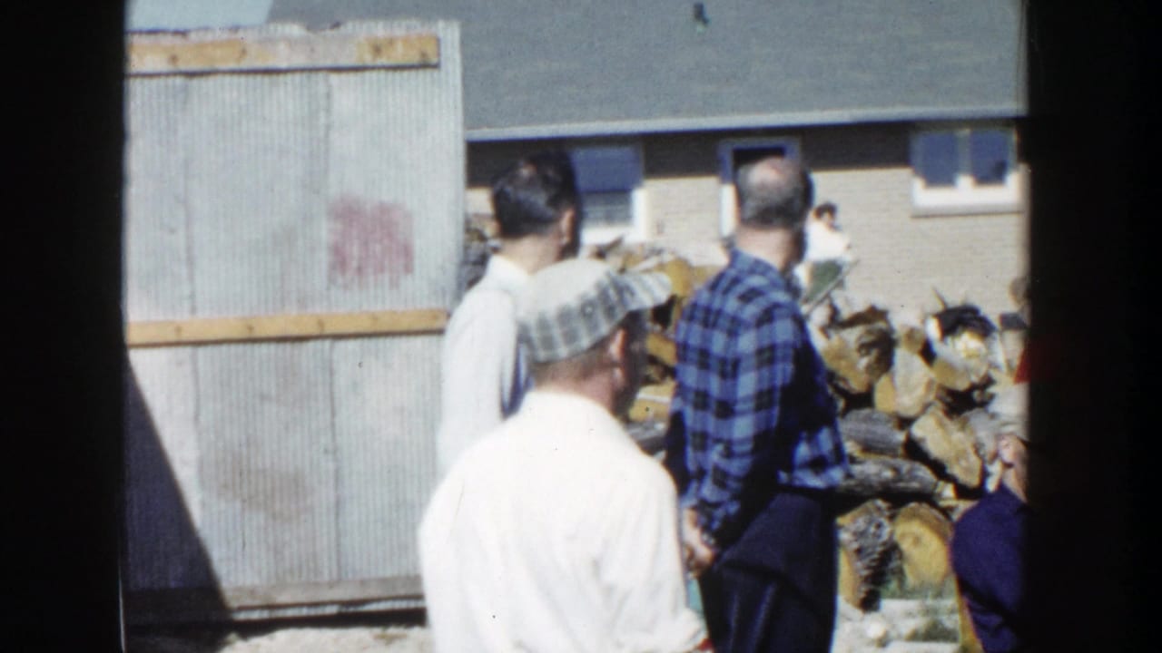 1958 Wisconsin backyard footage with adults viewed from behind near stacked firewood, a corrugated structure, and a brick home.