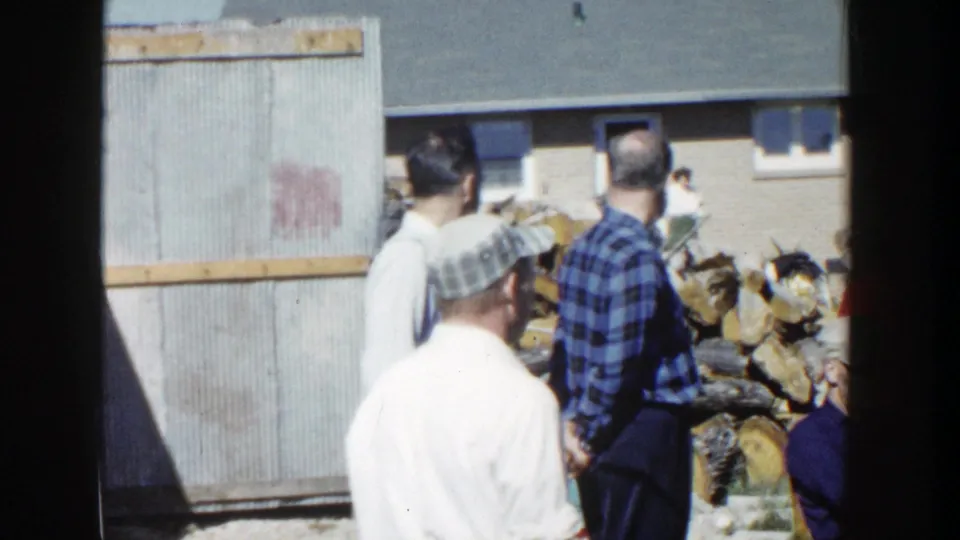 1958 Wisconsin backyard footage with adults viewed from behind near stacked firewood, a corrugated structure, and a brick home.