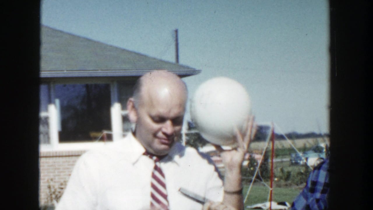 In 1958 Wisconsin home-movie footage, two adult men stand outdoors as one raises a large white sphere near a low suburban house.