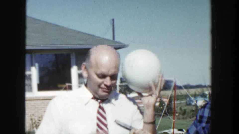 In 1958 Wisconsin home-movie footage, two adult men stand outdoors as one raises a large white sphere near a low suburban house.