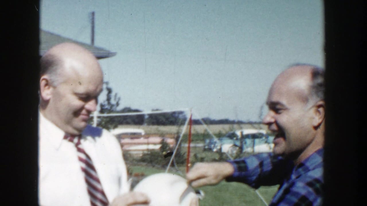 A man in a white shirt and striped tie smiles as a second man in a blue plaid shirt reaches toward the white sphere outdoors.