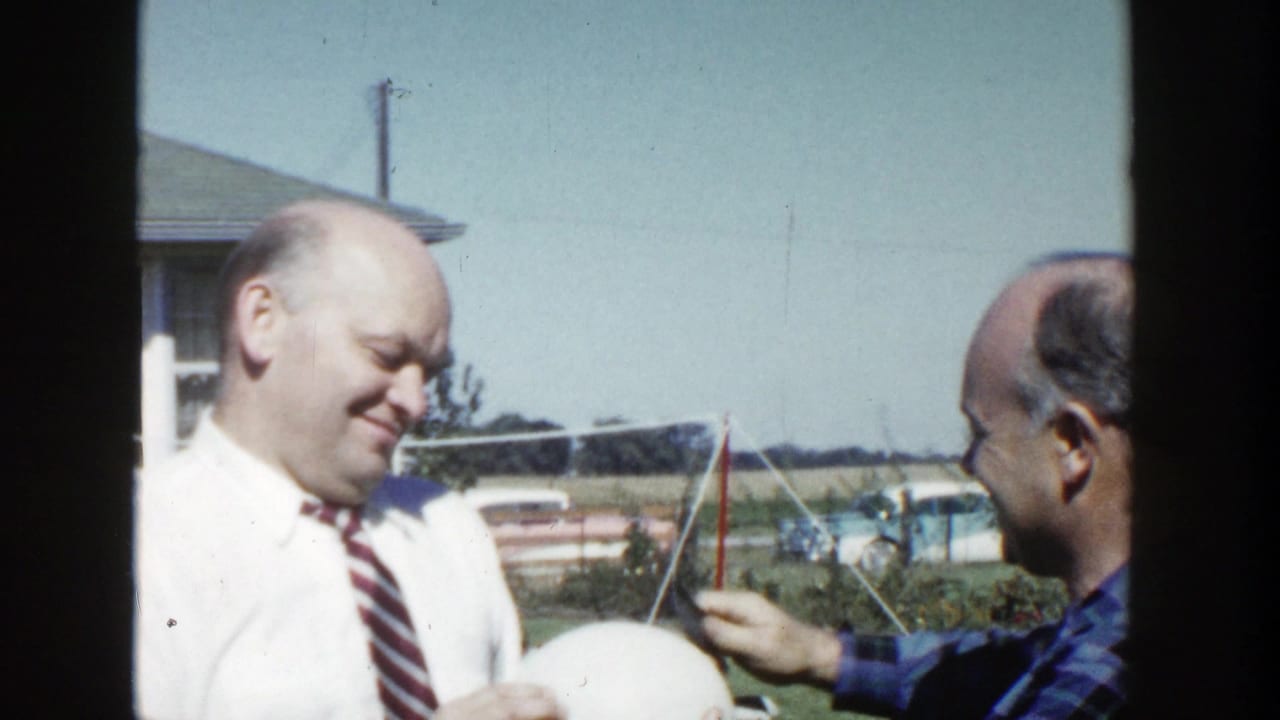 The two adult men face one another in the Wisconsin backyard, with the white sphere low in frame and cars visible beyond the lawn.