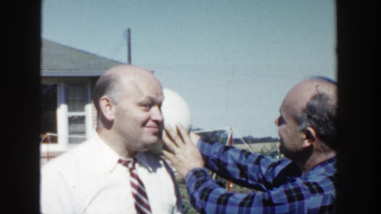 The man in the blue plaid shirt holds the white sphere close to the smiling man in a white shirt and striped tie outdoors.