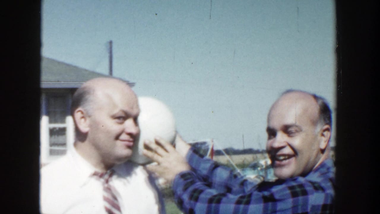 In 1958 Wisconsin home-movie footage, a man in a blue plaid shirt holds a white ball near a man wearing a white shirt and striped tie.
