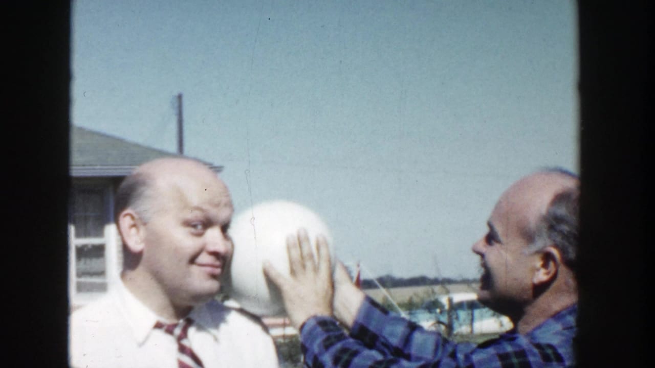 The plaid-shirted man holds the white ball against the side of the other man's head in a sunny Wisconsin yard.