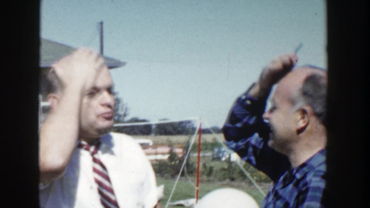The plaid-shirted man appears to pretend to comb or groom the other man's head as the man in the tie makes a comic expression.