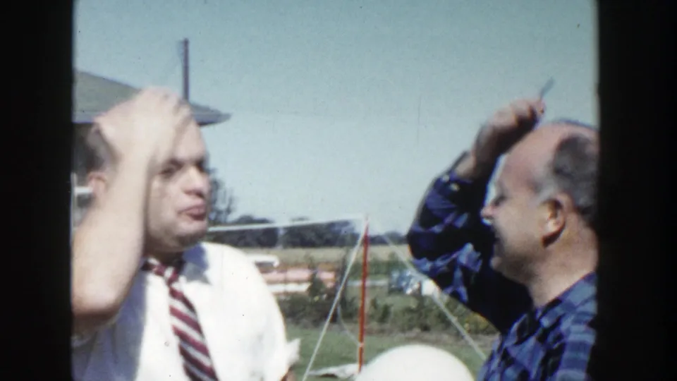 The plaid-shirted man appears to pretend to comb or groom the other man's head as the man in the tie makes a comic expression.
