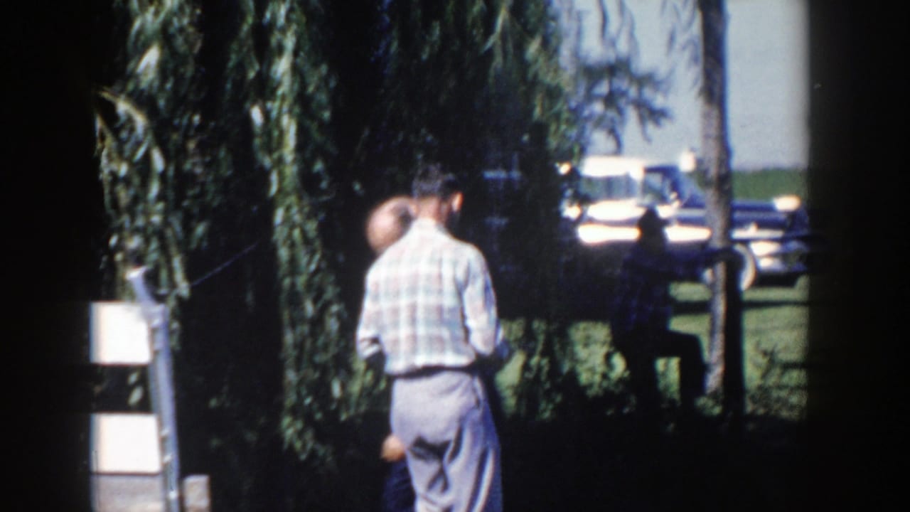 Two adult men in 1950s attire stand outdoors near a weeping willow, with a parked car beyond the grass.