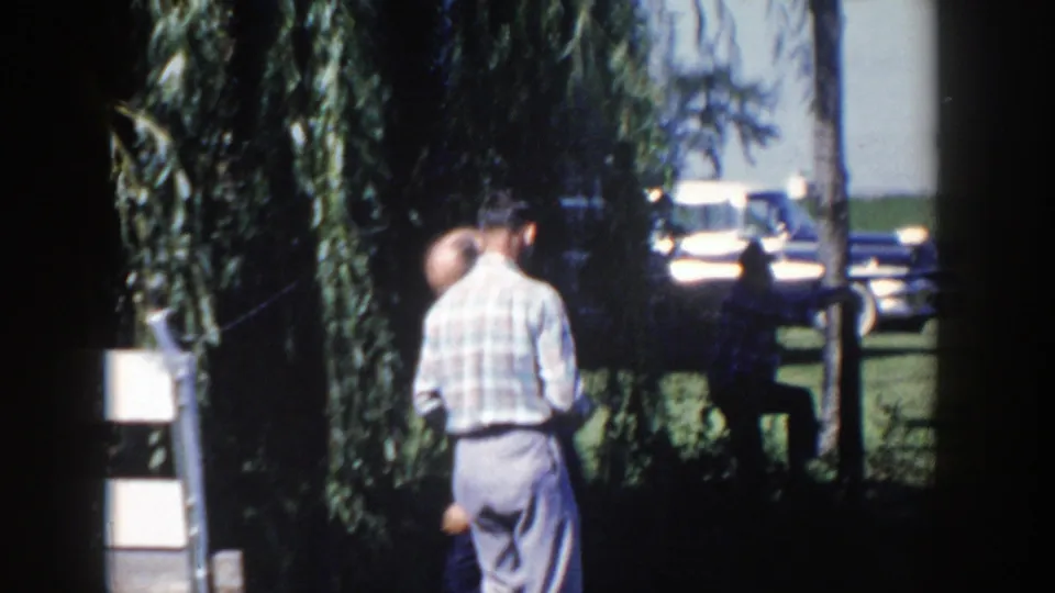 Two adult men in 1950s attire stand outdoors near a weeping willow, with a parked car beyond the grass.