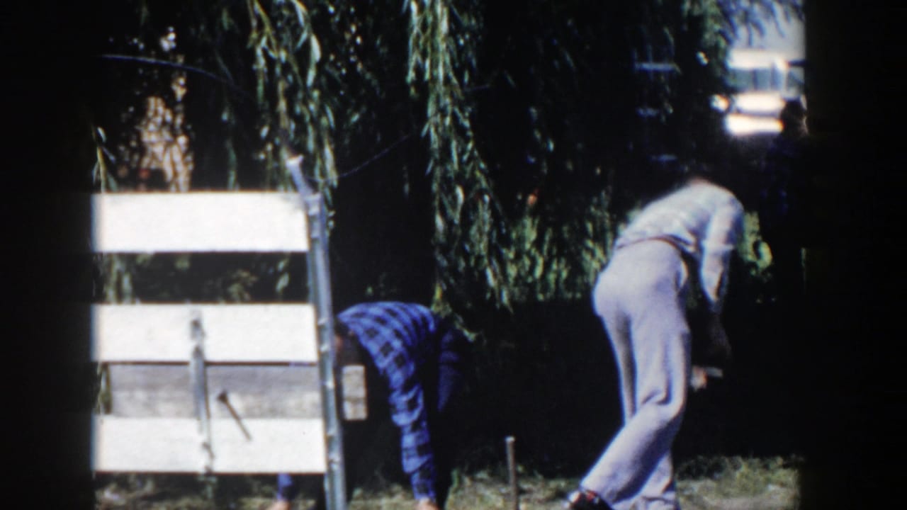 In the Wisconsin yard scene, two men bend down beside pale boards or boxes, handling small objects on the grass.