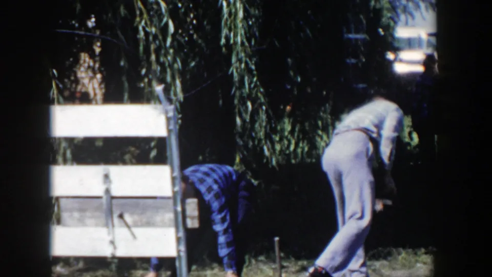 In the Wisconsin yard scene, two men bend down beside pale boards or boxes, handling small objects on the grass.