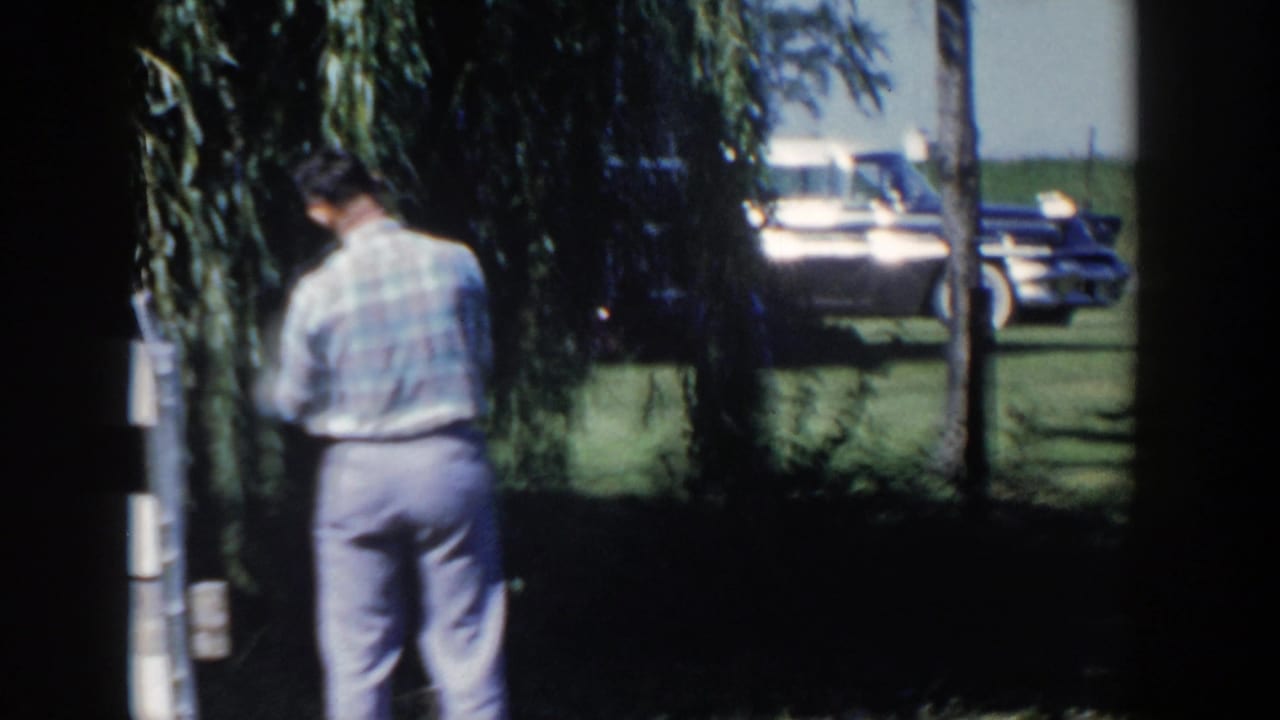 In 1958 Wisconsin home-movie footage, a man in a plaid shirt stands near a willow tree as cars sit in the background.