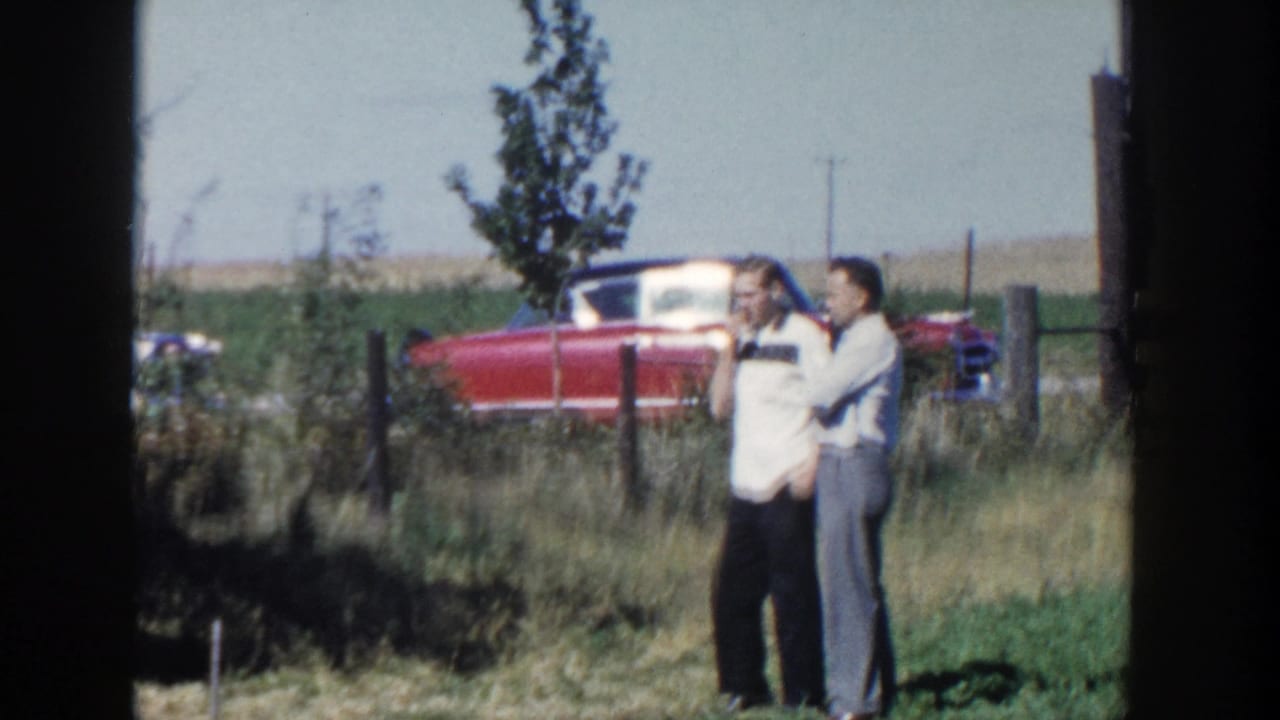 Two adult men stand together in a Wisconsin field, with a red classic car and fence line behind them.