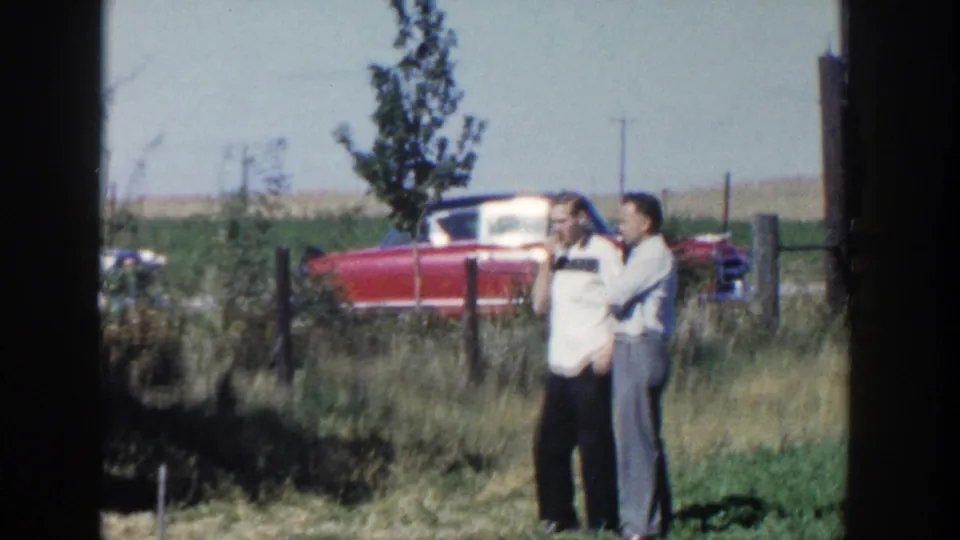 Two adult men stand together in a Wisconsin field, with a red classic car and fence line behind them.
