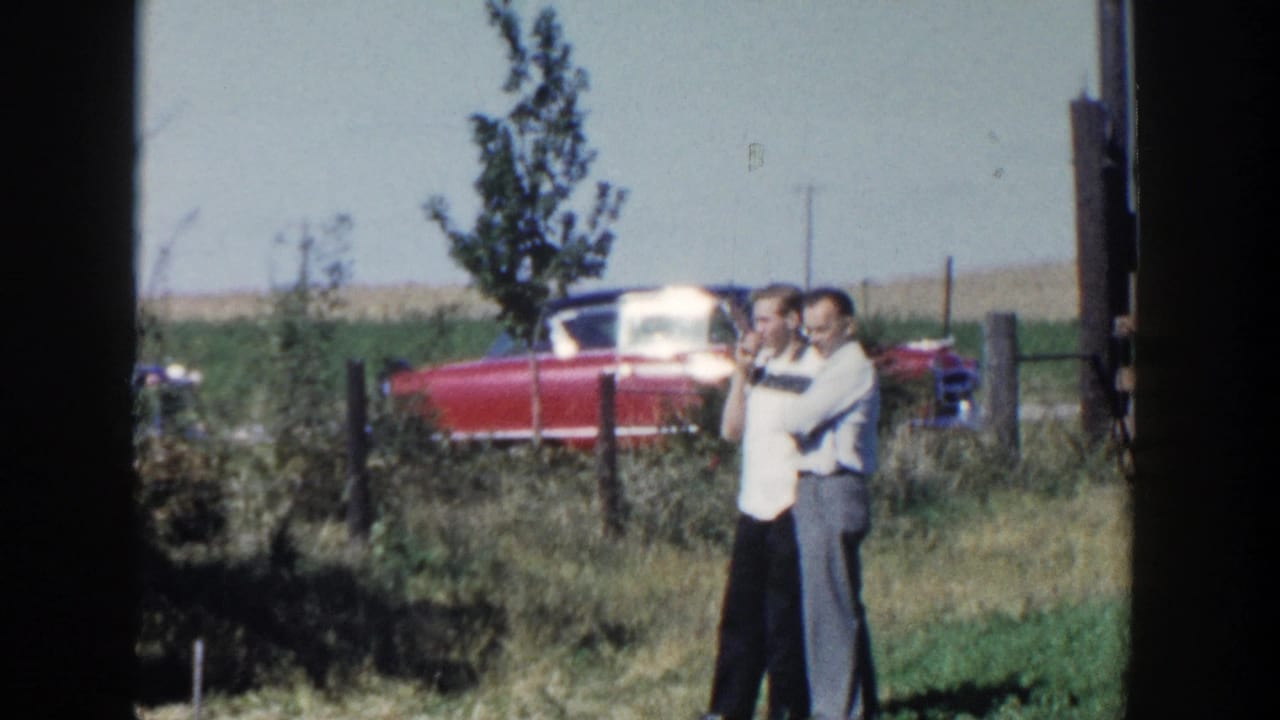 The two men remain near the fence in rural Wisconsin, one in a light shirt and the other in darker trousers, with the red car behind them.