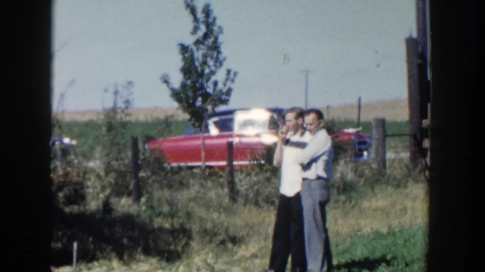 The two men remain near the fence in rural Wisconsin, one in a light shirt and the other in darker trousers, with the red car behind them.