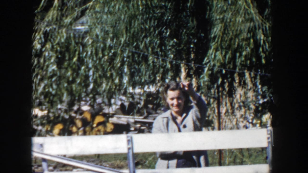 In 1958 Wisconsin home-movie footage, a woman in a grey jacket stands behind a white fence with willow branches and firewood behind her.