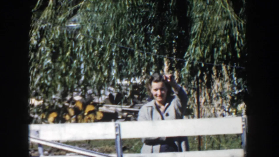 In 1958 Wisconsin home-movie footage, a woman in a grey jacket stands behind a white fence with willow branches and firewood behind her.