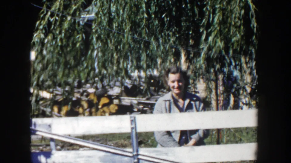 The woman remains behind the fence in the Wisconsin backyard, smiling toward the home-movie camera beneath hanging willow branches.