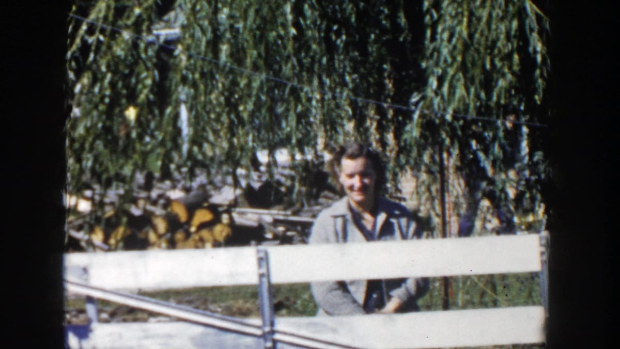 A 1958 Wisconsin home-movie frame shows the woman behind white fence rails, with soft focus, strong sunlight, trees, and stacked firewood nearby.