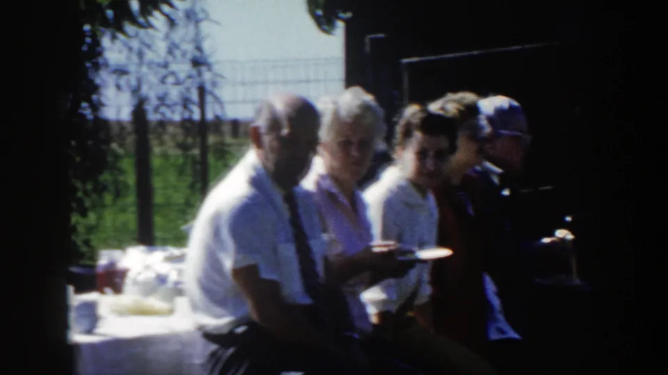 1958 Wisconsin home-movie frame of adults seated in shade with plates near a table, fence, grass, and trees in the background.