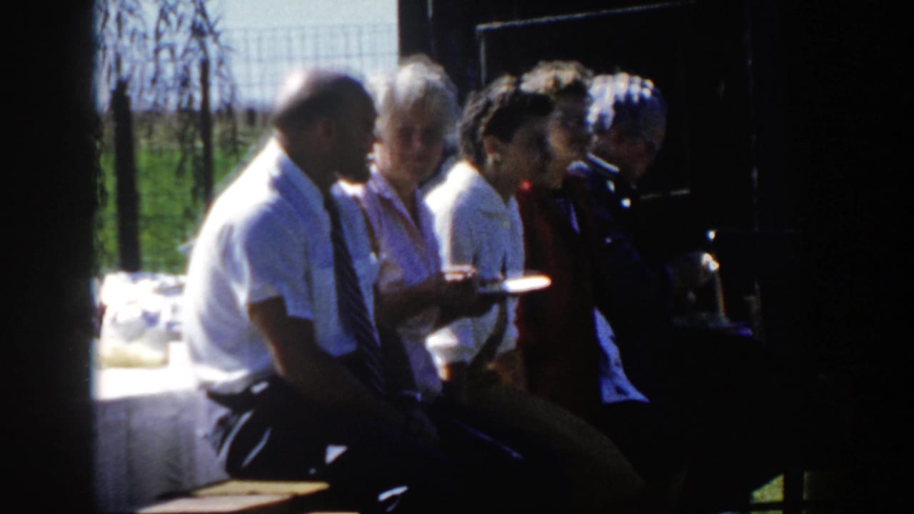 1958 Wisconsin home-movie frame showing adults seated shoulder to shoulder with plates beside a sunlit table and wire fence.