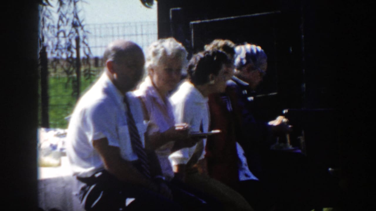 1958 Wisconsin home-movie frame of adults eating or waiting with plates near a table, with bright grass and fencing behind them.