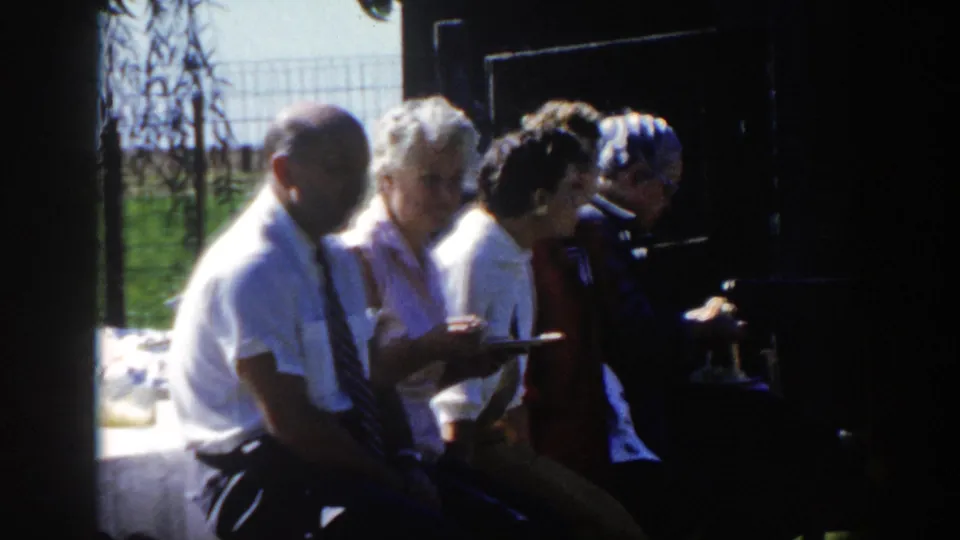 1958 Wisconsin home-movie frame of adults eating or waiting with plates near a table, with bright grass and fencing behind them.