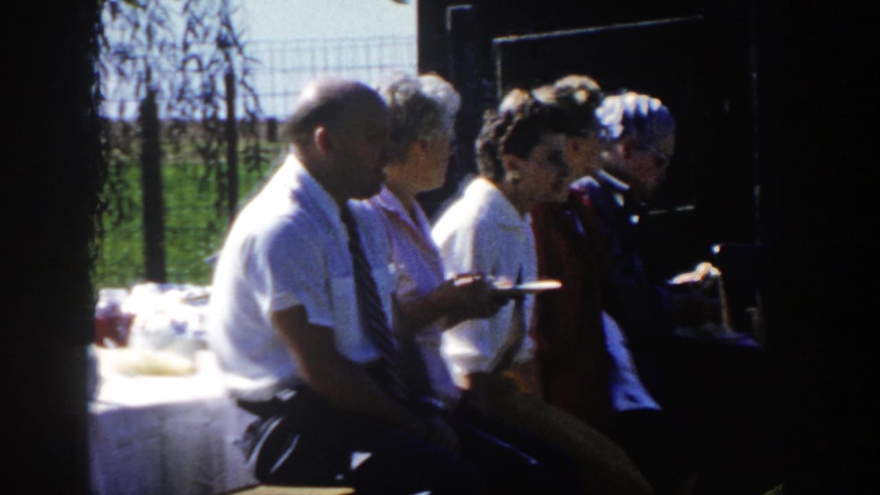 1958 Wisconsin home-movie frame of seated adults in profile holding plates in shade, with a serving table and fenced greenery behind.