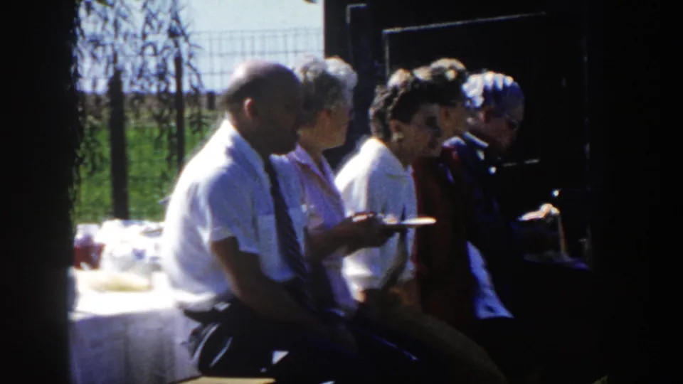 1958 Wisconsin home-movie frame of seated adults in profile holding plates in shade, with a serving table and fenced greenery behind.