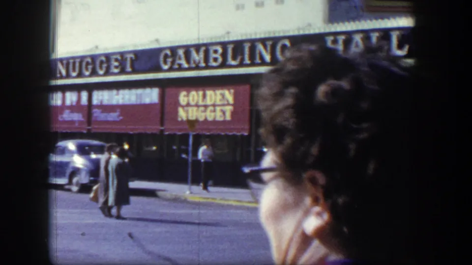 1954 Las Vegas Golden Nugget street scene with pedestrians, parked cars, and a close foreground bystander partly blocking the view.